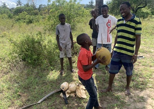 Local Kenyan kids and adults in Mida Creek stand under a coconut tree as a child holds a large coconut with two hands.