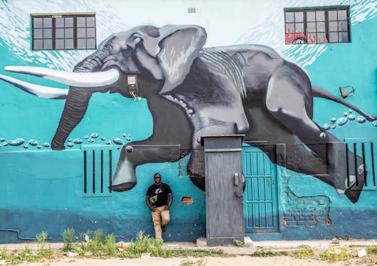 A street art mural showing a giant elephant jumping, with its tusks pointed to the left, on an aquamarine blue background. An adult Black man is standing against the wall in long khaki pants and a black t shirt with one leg up against the wall.