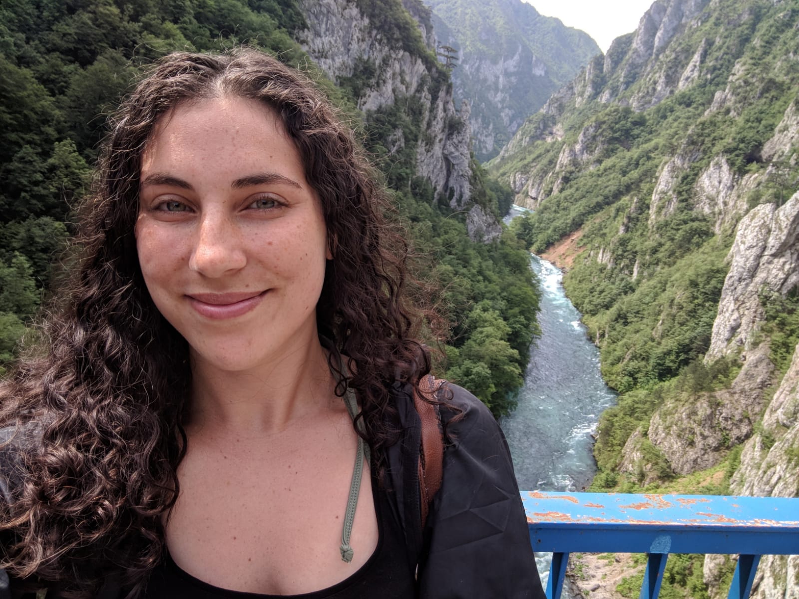 A headshot of a young Caucasian woman with long, brown curly hair standing on a bridge with a blue railing over a gorge with a river running between a rocky canyon.