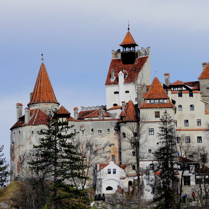 Bran Castle, a blocky beige building with pointed red brick rooftops, sits majestically against a cloudy blue sky with dark thin trees in the foreground.
