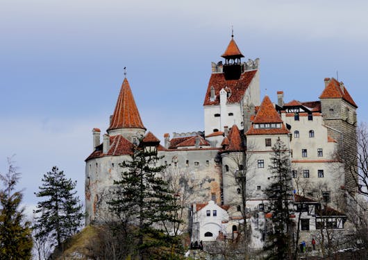 Bran Castle, a blocky beige building with pointed red brick rooftops, sits majestically against a cloudy blue sky with dark thin trees in the foreground.