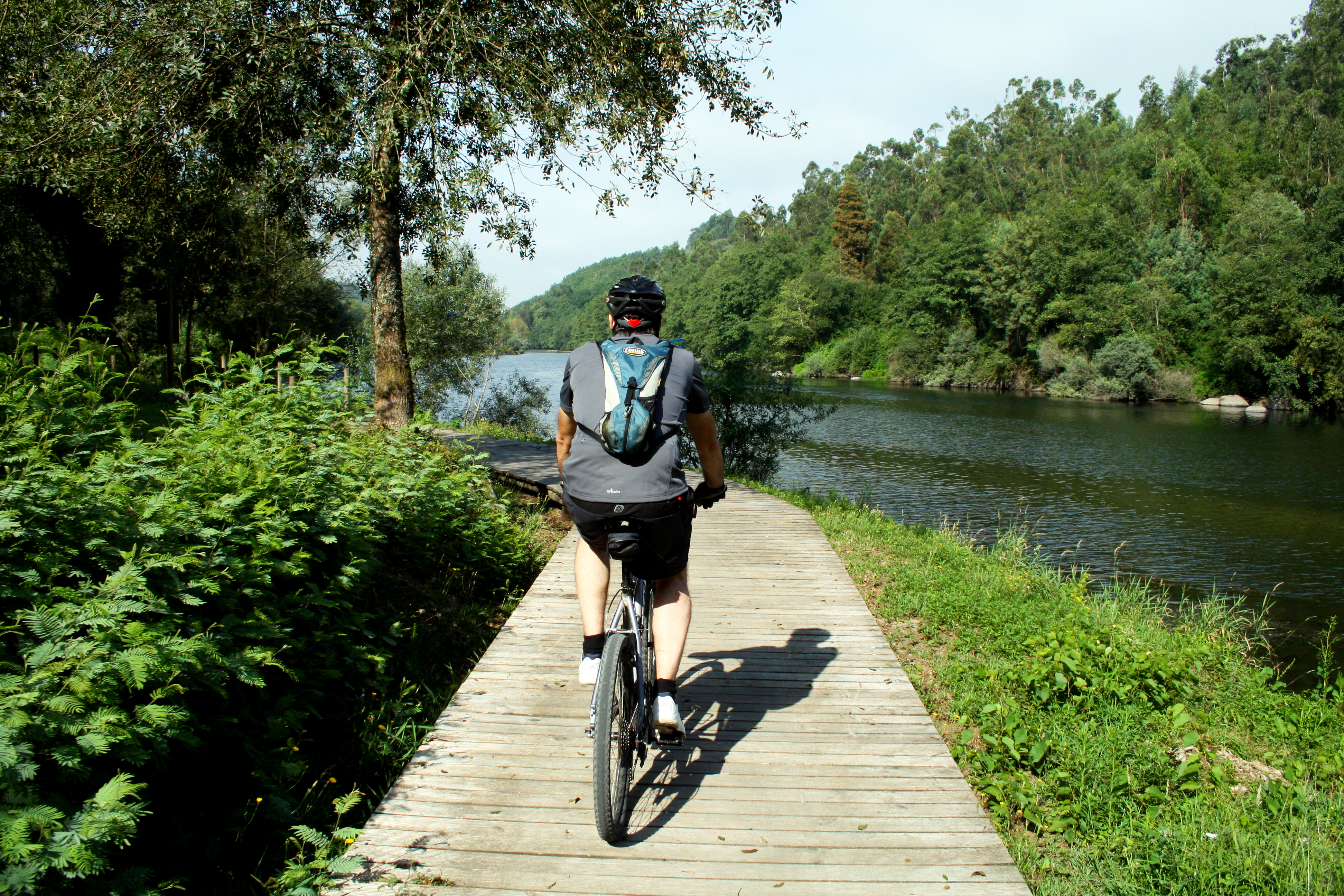 An adult Caucasian man bikes along a boardwalk with lush greenery to his left and a flowing river to his right in Porto's Alto Minho region.