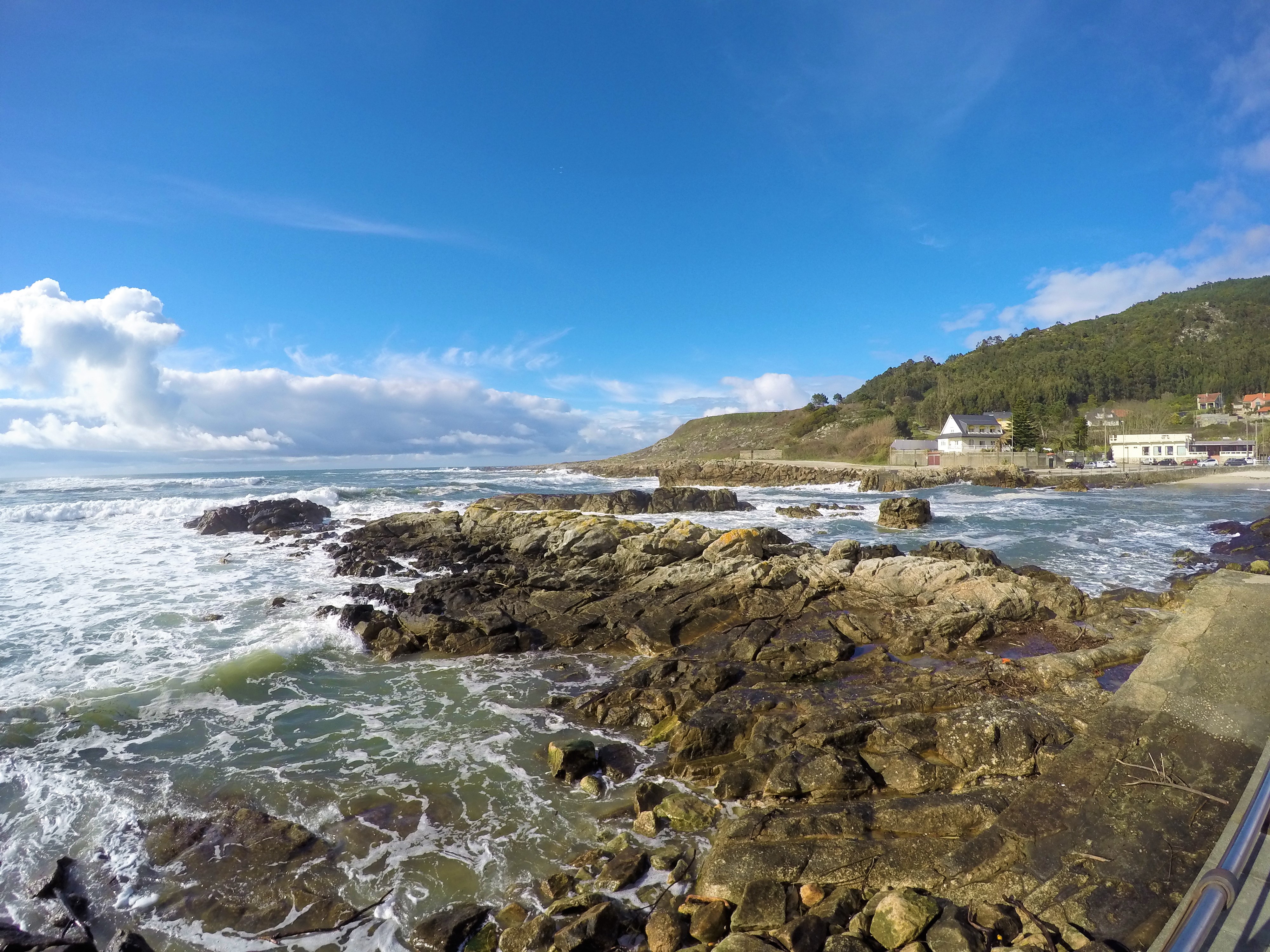 Waves crash up against a rocky shore in norther Portugal along the Coastal Camino de Santiago, under a blue sky with a few white clouds on the horizon.