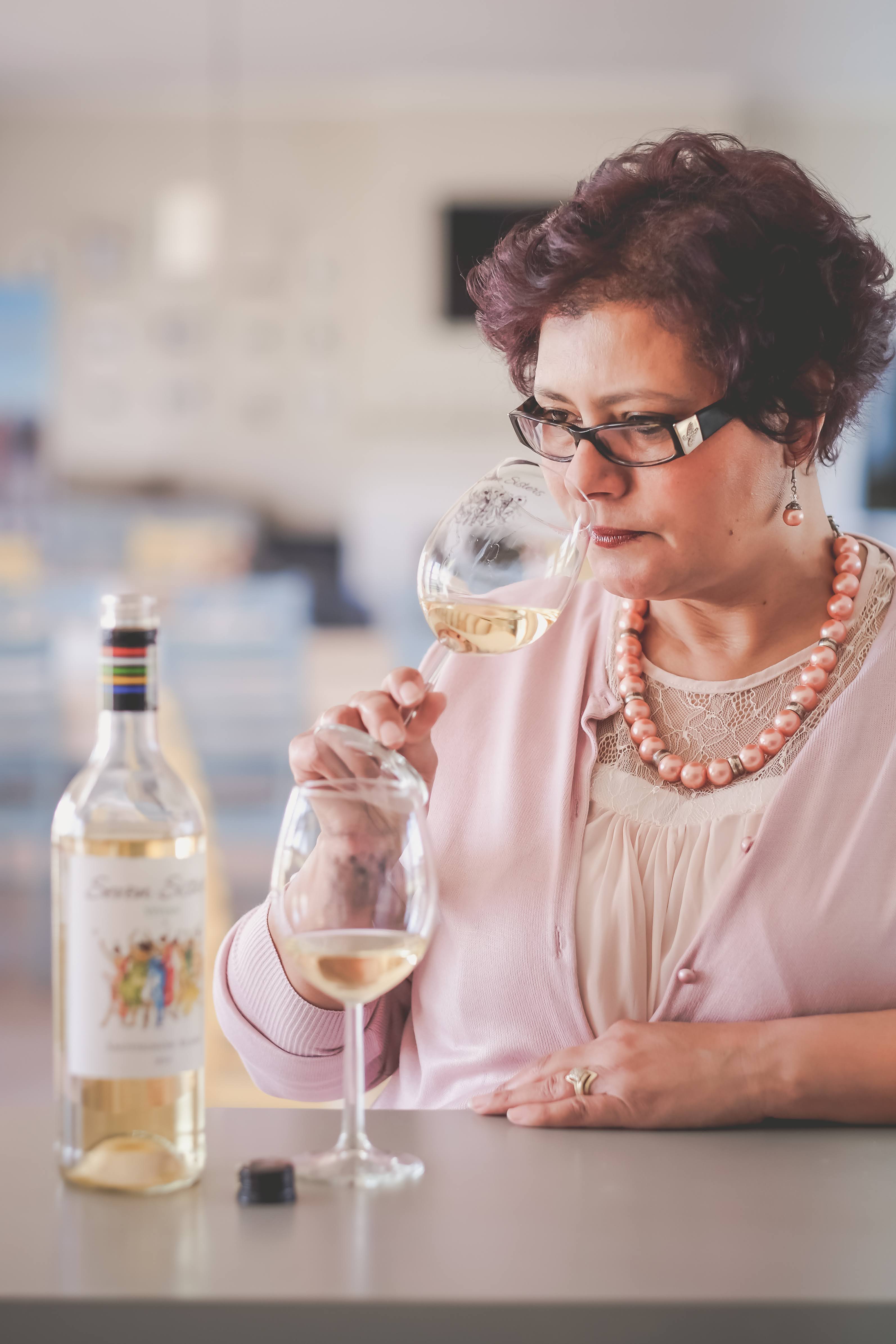 A Caucasian woman is smelling a glass of white wine with a bottle and another glass on the counter. The woman is middle-aged, she has short, brown hair, and glasses. She is wearing a blouse, pearls, a sweater, and a pearl necklace, all different tones of pink. The woman is in an indoor environment. 