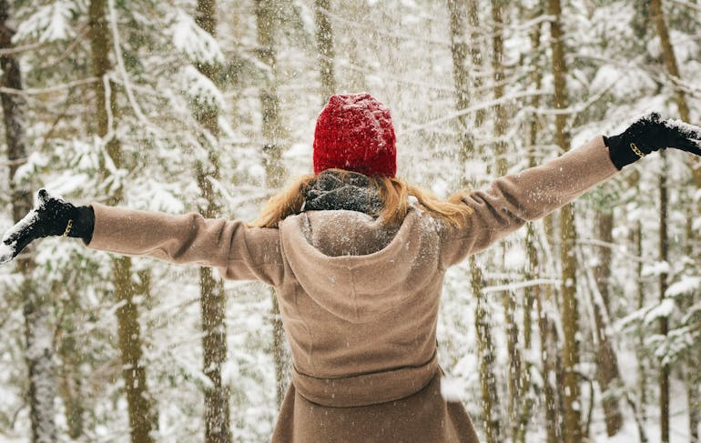 A woman with long, straight blond hair wearing a beige coat, red had, and black gloves stands with her back to the frame with her arms outstretched, forming a T, amidst a snowy forest of thin trees.