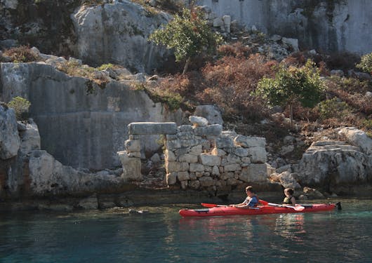 A white man and woman sit in a red double kayak with their paddles horizontal above the water, with ancient Roman ruins in the background.