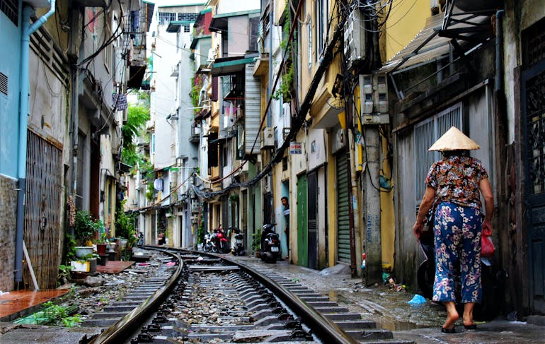 A woman in blue floral pants and darker floral short sleeve shirt and a traditional Vietnamese sunhat walks to the right side of a railroad traffic through a narrow street full of multi-level colorful houses.
