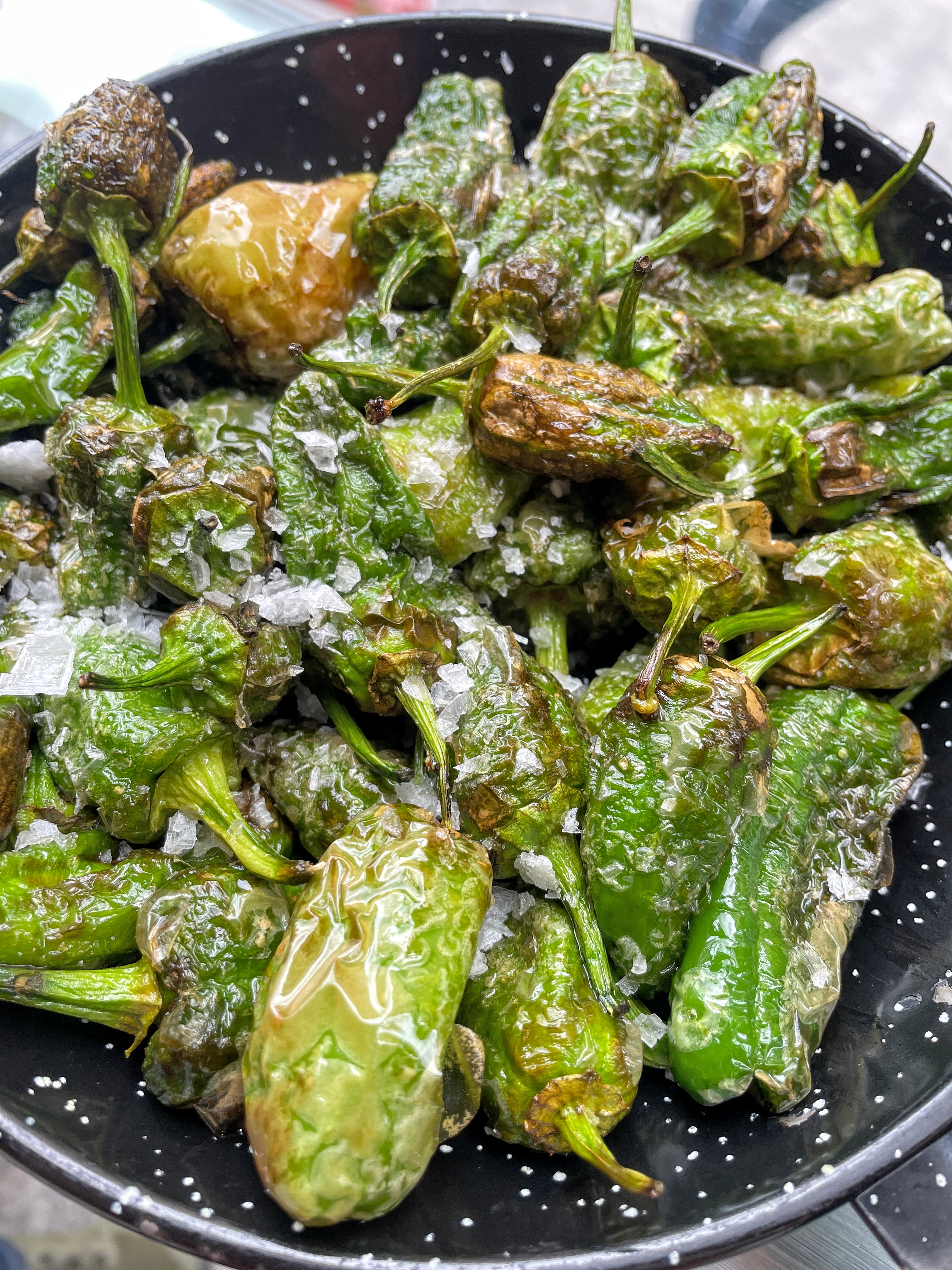 A plate of bright green, blistered peppers - pimientos de Padron - covered in salt are served from a warm skillet.
