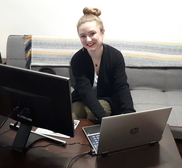 Caucasian woman sits on a grey couch, looking up smiling, while facing an open laptop computer and a monitor screen set up on a brown wooden table.