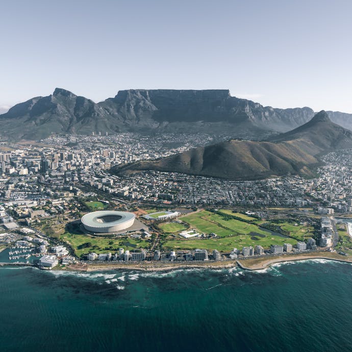 An aerial view from the water of Cape Town, South Africa, showing the bird's nest athletic stadium in the foreground and the flat-topped Table Mountain in the background. Slow Travel through South Africa on this Wellness, Wine, & Safari tour.