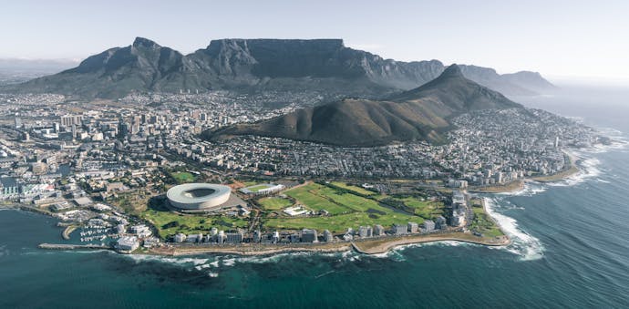 An aerial view from the water of Cape Town, South Africa, showing the bird's nest athletic stadium in the foreground and the flat-topped Table Mountain in the background. Slow Travel through South Africa on this Wellness, Wine, & Safari tour.