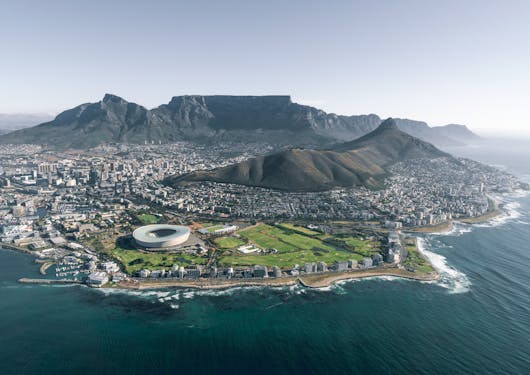 An aerial view from the water of Cape Town, South Africa, showing the bird's nest athletic stadium in the foreground and the flat-topped Table Mountain in the background. Slow Travel through South Africa on this Wellness, Wine, & Safari tour.