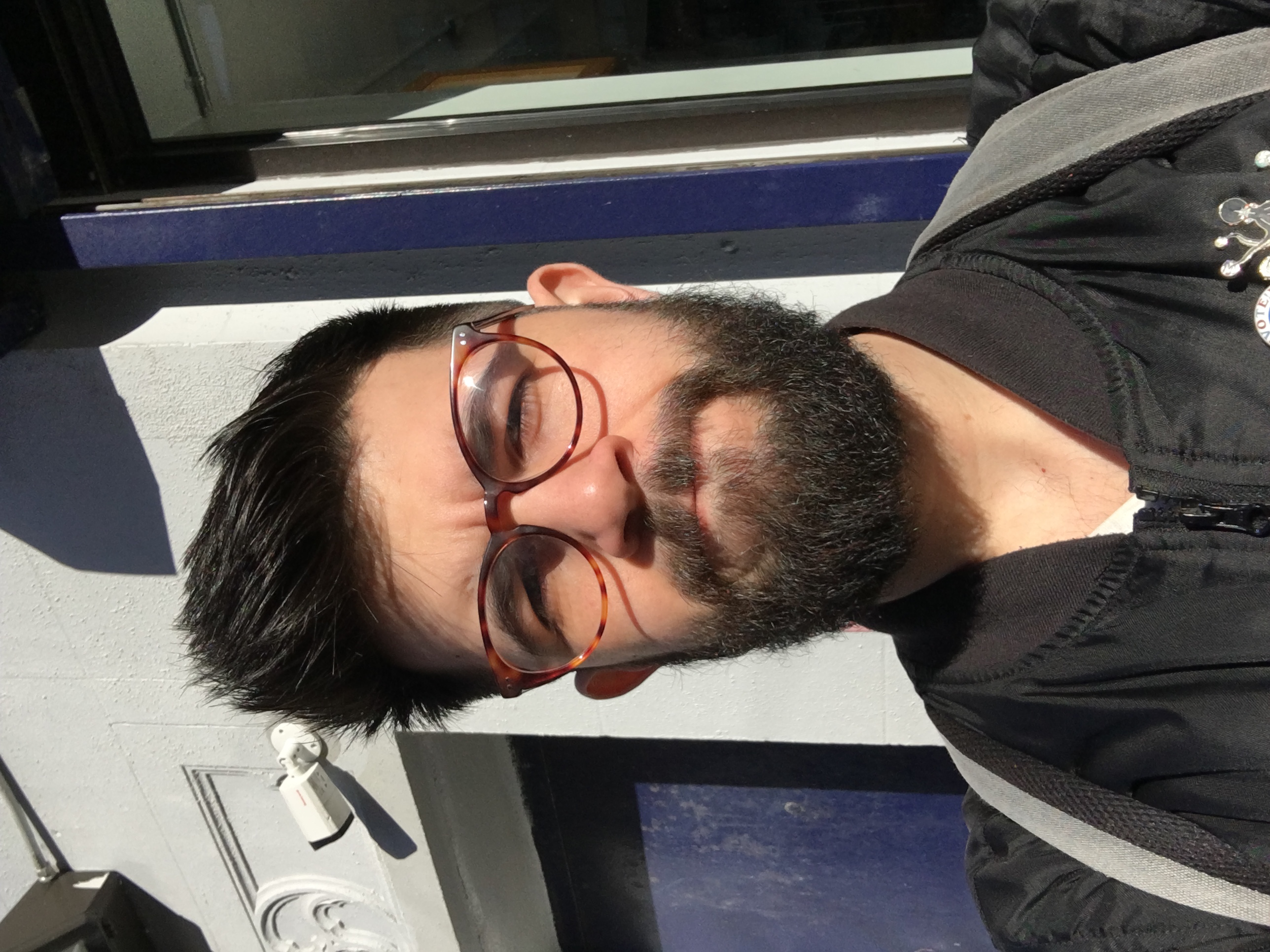 A headshot of a young Caucasian man in a black zip up jacket, dark short hair, red glasses, and a dark beard, looking at the camera standing in front of a beige and blue building.