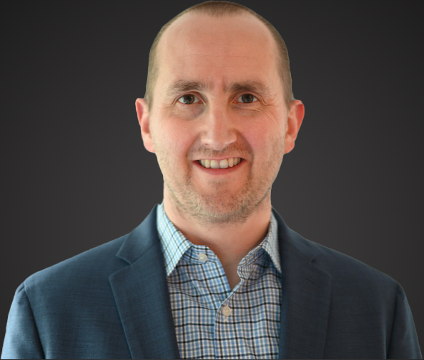 A headshot of a young Caucasian man with a buzz cut,a 5-o-clock shadow, and wearing a blue patterned button up shirt under a blue blazer, smiling at the camera.