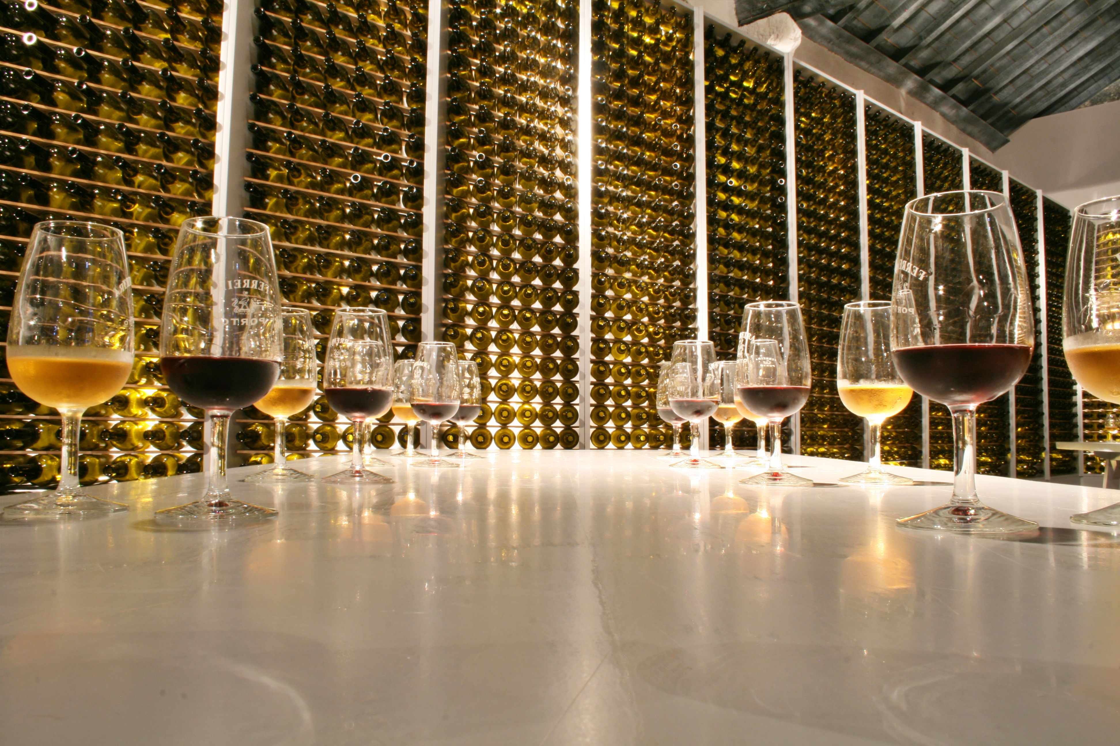 14 wine glasses in 7 sets of 2, with one of each glass containing red wine and the other containing white wine, form an almost V shape with the point at the far end of the frame. Behind the point is a floor to ceiling wine rack filled with bottled. Portugal.