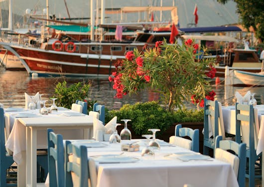 A restaurant terrace shows tables, with with white tablecloths and blue chairs, in front of a port or harbor with several boats in the background. This is at sunset along the Lycian Way in Turkiye.