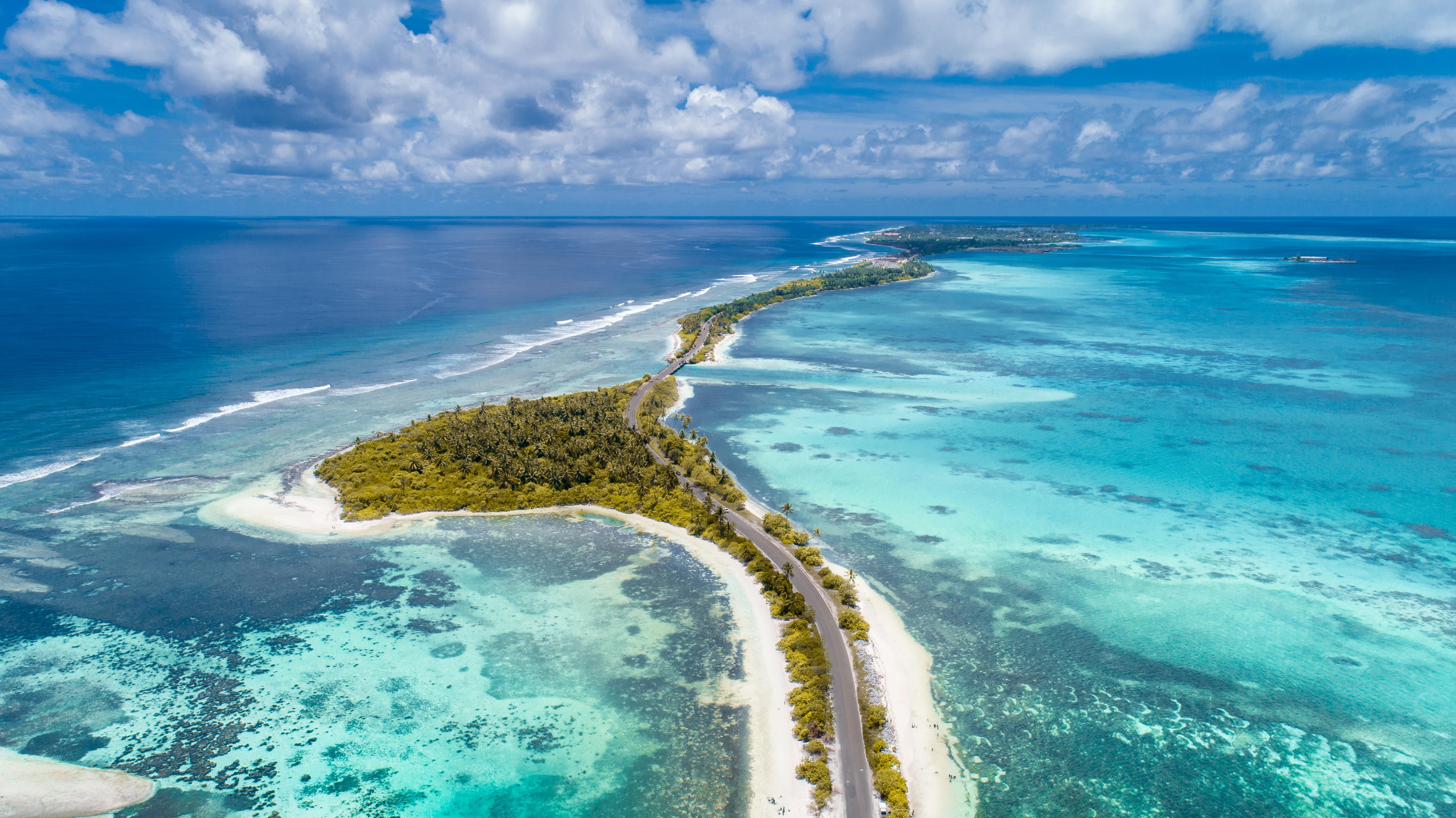Beautiful white sand beaches sprawl through sparkling shallow blue water in the Maldives; green trees cover the land area, except a small paved road cutting through along the coast.