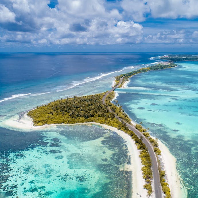 Beautiful white sand beaches sprawl through sparkling shallow blue water in the Maldives; green trees cover the land area, except a small paved road cutting through along the coast.