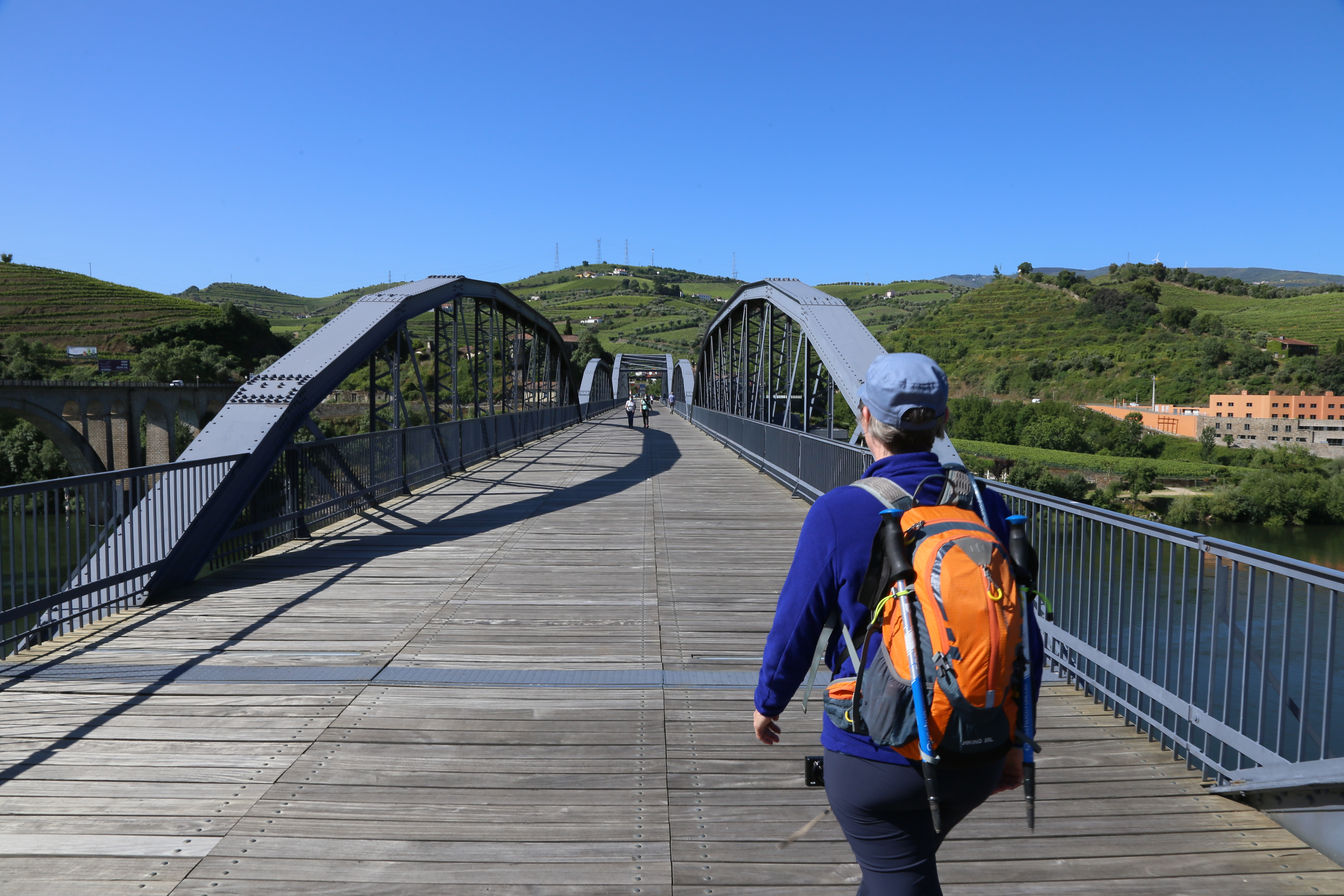 An adult, Caucasian female walker wearing a long sleeve blue pullover and a blue hat and an orange backpack with walking poles attached walks away from the camera over a bridge. Green terraced vineyards in Portugal's Douro region are on the other side.