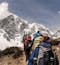 A group of climbers or trekkers carrying backpacks walk in a single file line on a packed dirt path towards snow-capped mountains.
