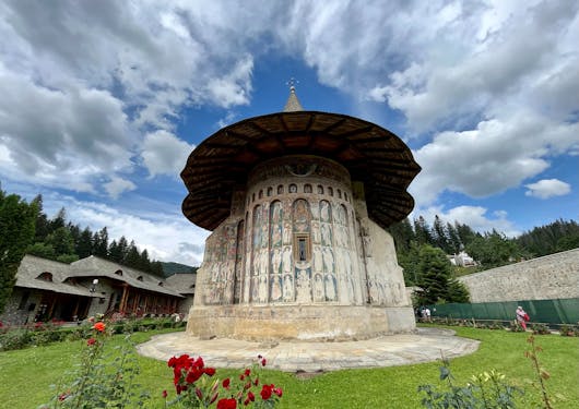 The painted monastery in Bucovina, Romania sits with a paved stone walkway around it and grass in the foreground. It's a cloudy day. The circular church's exterior is covered in painted depictions.