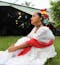 An indigenous woman from Mexico sits on green grass in a white dress with colorful flowers in her hair.