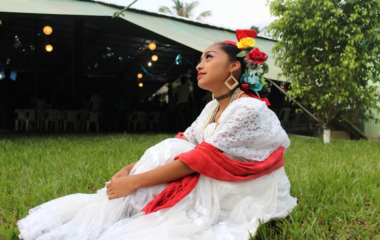 An indigenous woman from Mexico sits on green grass in a white dress with colorful flowers in her hair.