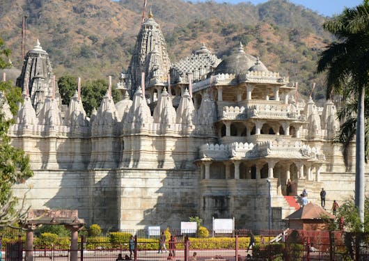 The Ranakapur Jain Temple is shown from a slight distance and is offset from center. You can see the beautiful and ornate architecture of the beige building with a brown and green hillsite behind it. There are a couple trees to the left and right of the frame, and you can see several people walking up the red lined steps leading into the temple.