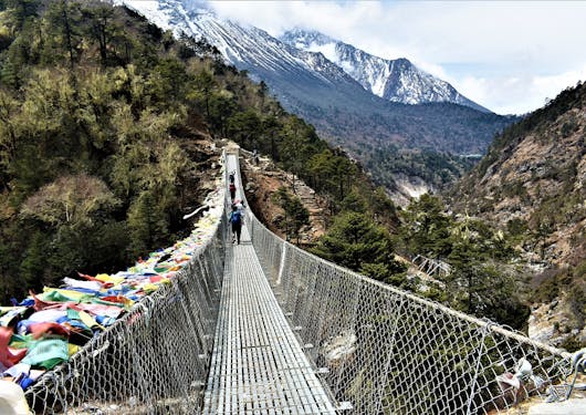 A chain link bridge with prayer flags along the lefthand side extends across a valley into a wooded area, with snow-capped mountains in the background.