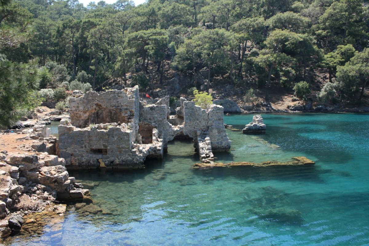 Clear, turquoise water is shallow enough where you can see sunken roman ruins below, with a rocky shore to the left and green trees in the background.