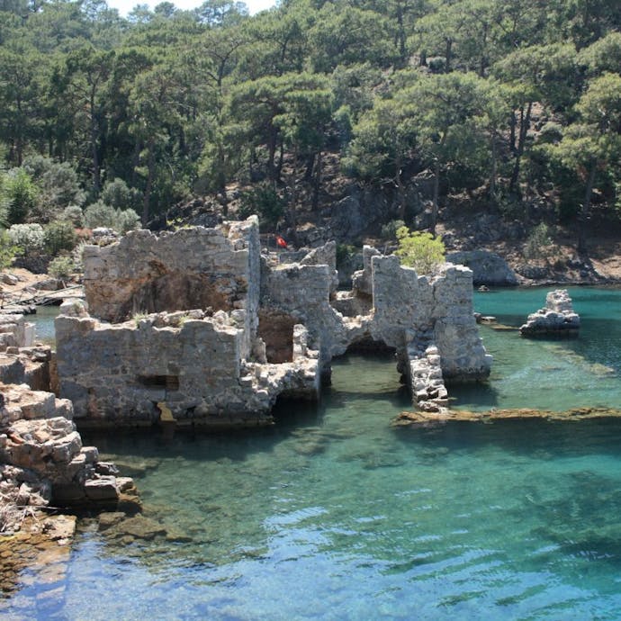 Clear, turquoise water is shallow enough where you can see sunken roman ruins below, with a rocky shore to the left and green trees in the background.