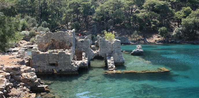Clear, turquoise water is shallow enough where you can see sunken roman ruins below, with a rocky shore to the left and green trees in the background.