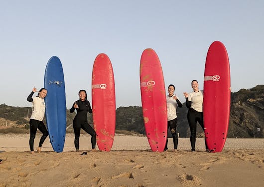 4 women - 3 Caucasian and 1 Asian - stand with their surfboards. 3 surfboards are red and one is blue. They are smiling and looking at the camera, standing on the sandy beach with a blue sky behind them.