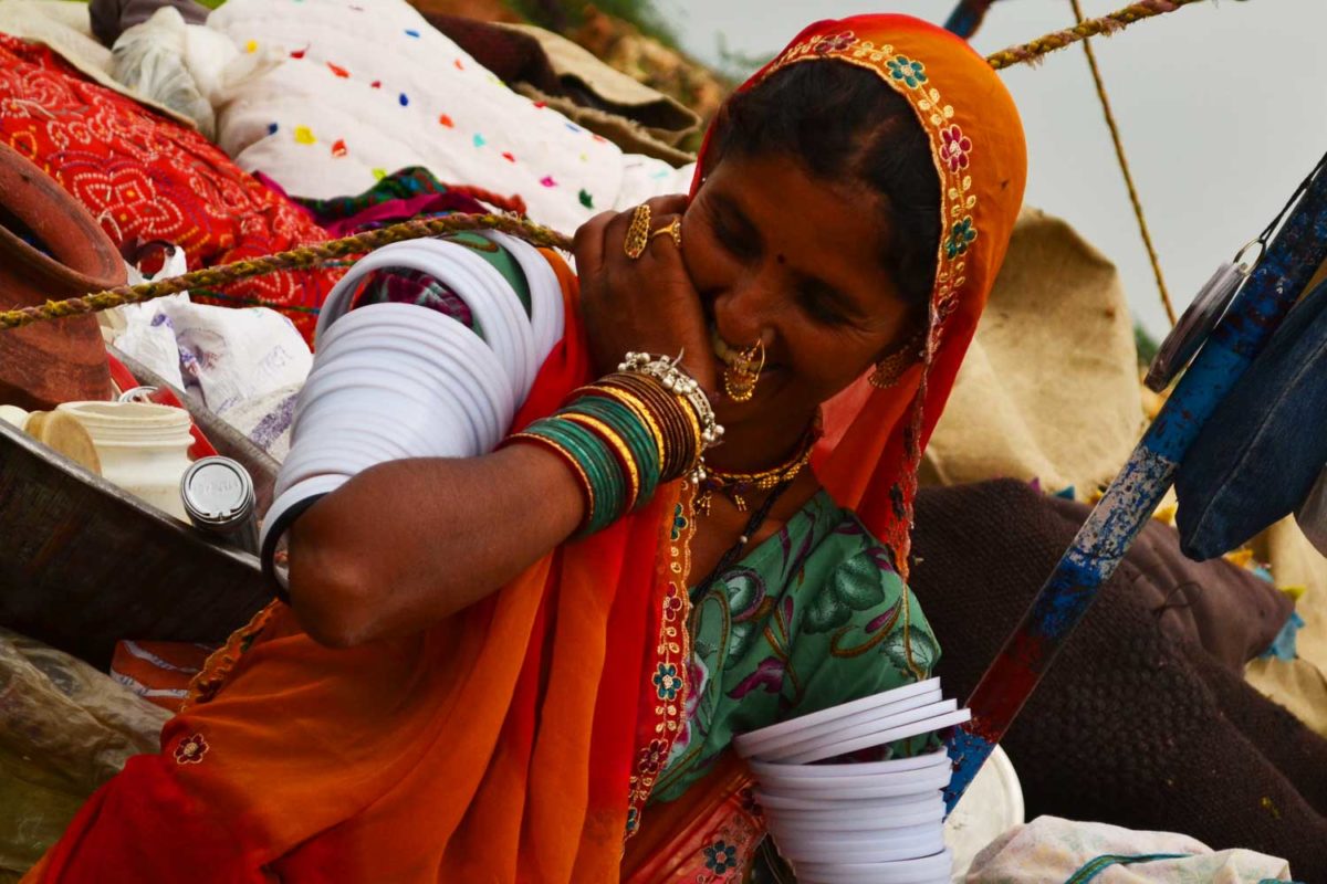 A local Indian woman in the traditional garb of a red and green sari wrap and head cover and lots of bangles and jewelry smiles into her hand shyly, not looking at the camera.