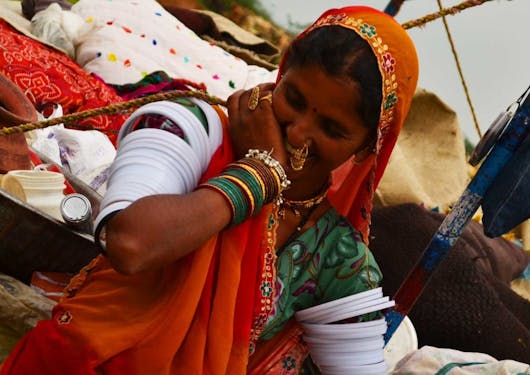 A local Indian woman in the traditional garb of a red and green sari wrap and head cover and lots of bangles and jewelry smiles into her hand shyly, not looking at the camera.