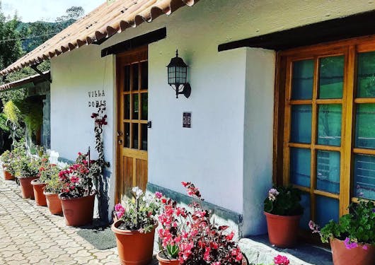 The front facade of Villa Doris Country Lodge, an eco-friendly, sustainable lodge in Ecuador is shown with white-washed walls and colorful potted flowers in a line in front of the building. Wood-paned windows and a wood-paned door are also shown.