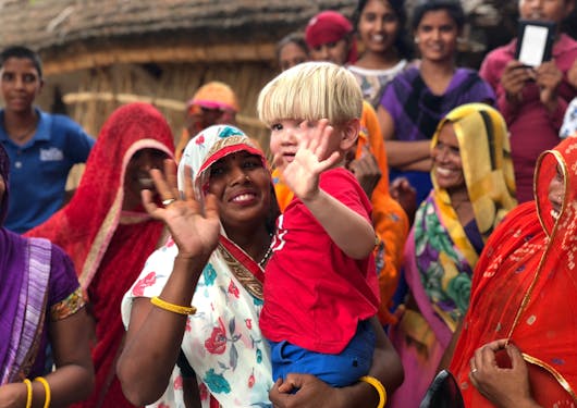 A group of smiling Indians all in colorful traditional dress are shown on the Golden Triangle Cultural Tour. An Indian woman holds a white, young, blond boy and they both have one hand raised waiving to the camera; they are both smiling.