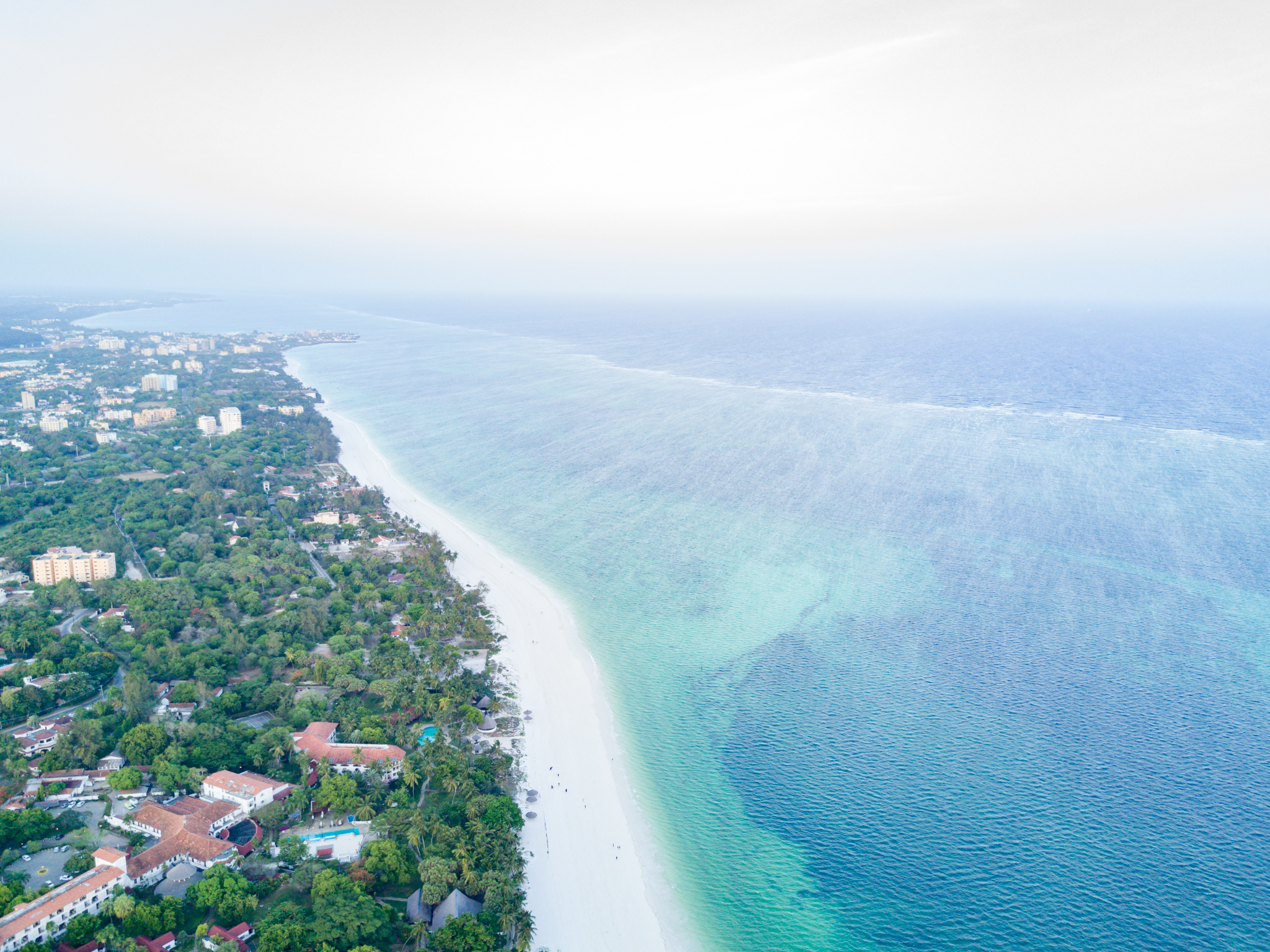 The Kenyan coast is shown from an aerial view with the land on the left with green trees and some buildings, a white sand beach, and then the turquoise blue water to the right.