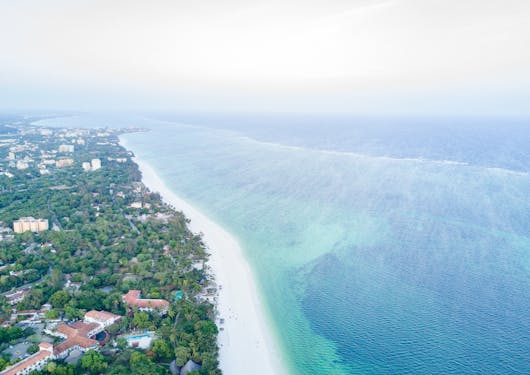The Kenyan coast is shown from an aerial view with the land on the left with green trees and some buildings, a white sand beach, and then the turquoise blue water to the right.