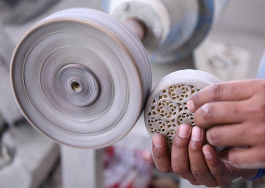 An Indian person shows their craft as a marble mason by holding up a circular piece of carved marble up to a buffer to smooth out the outside of it.
