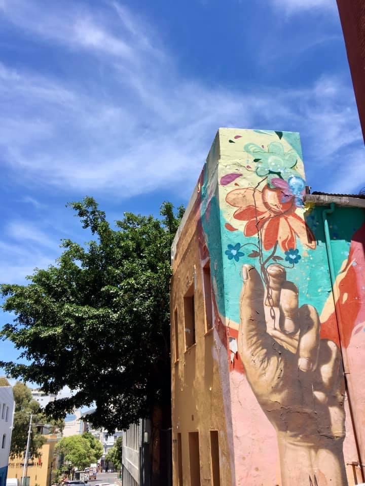 A colorful street mural on the side of a building shows a hand reaching for a flower. There is a green tree in the background and a blue sky above with some light wispy clouds.
