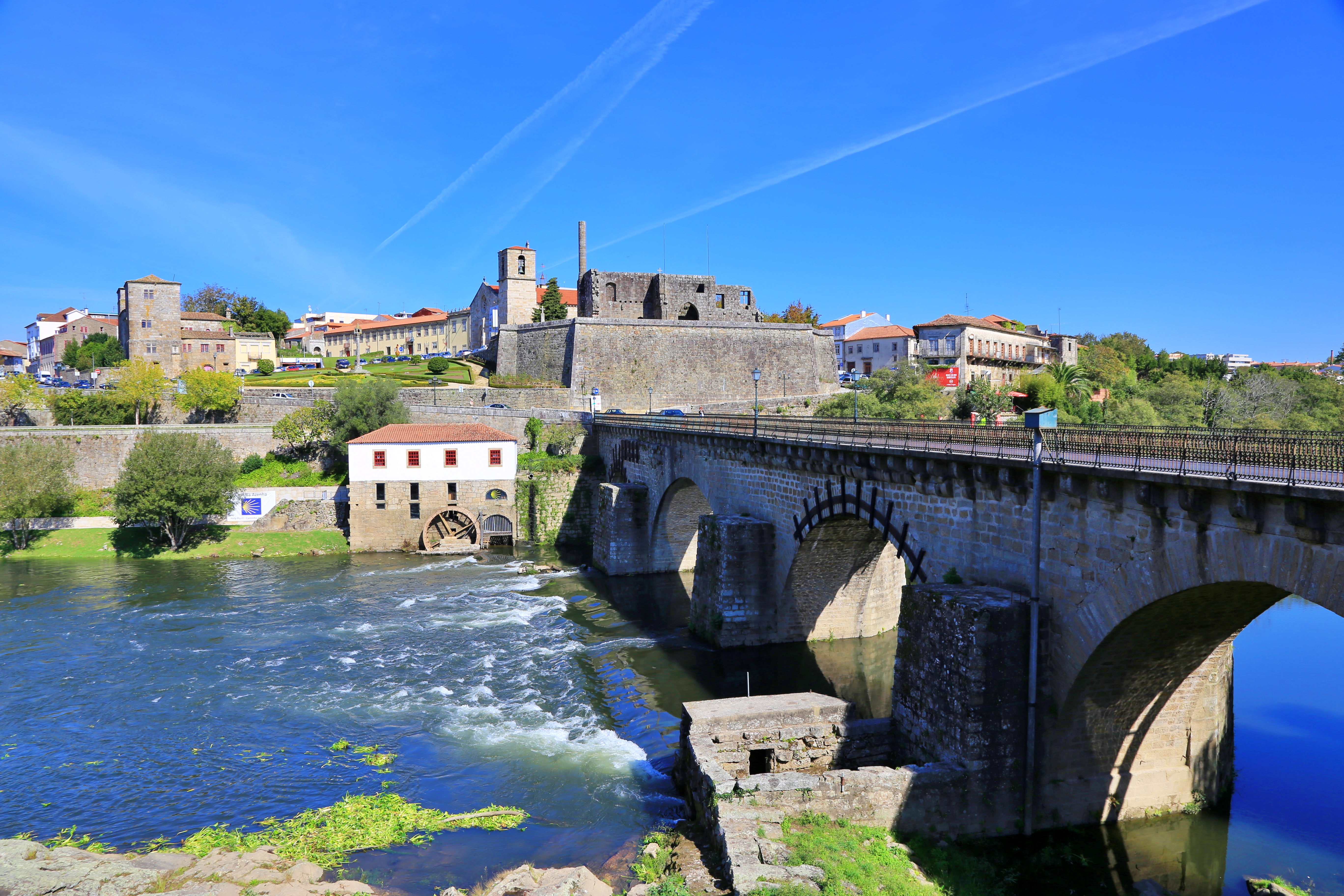 A sturdy stone bridge with three arches underneath spans a river leading into a fortified town in Portugal's Alto Minho region. The sky is blue and it's a sunny day.
