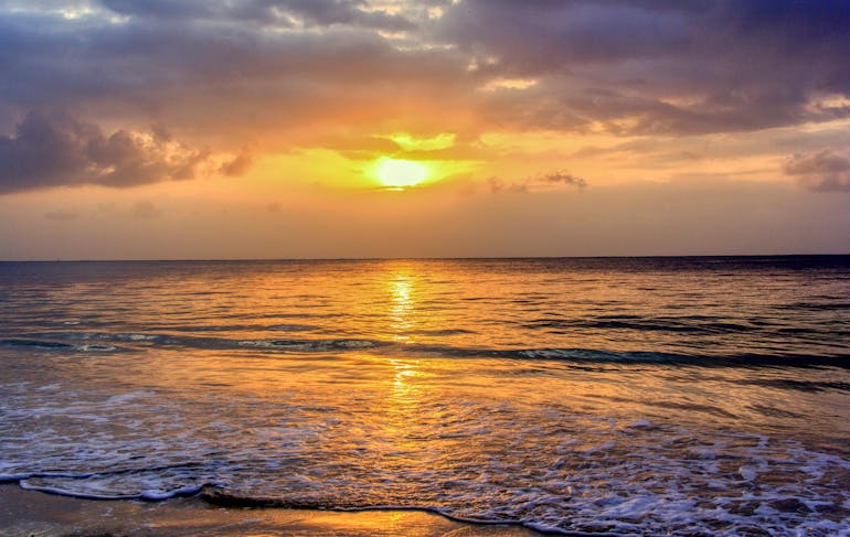 A stunning sunset off the Kenyan Coast is shown with a bright orange-yellow sun blasting through gray clouds and onto the shallow water.