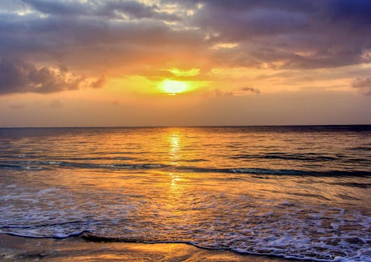 A stunning sunset off the Kenyan Coast is shown with a bright orange-yellow sun blasting through gray clouds and onto the shallow water.