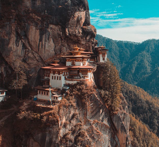The iconic Tiger's Nest Monastery near Paro, Bhutan sits nestled amongst steep cliffs, with forest in the background and a blue sky overhead.