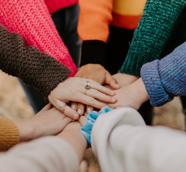 A group of 7 hands come together in the middle of the screen, one on top of the other; the long sleeves on the arms reflecting colors of red, orange, green, blue, brown, and beige.