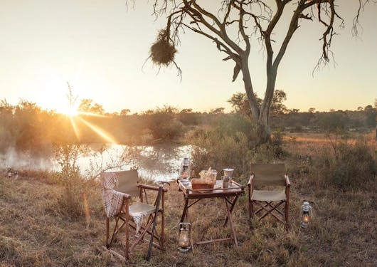 A romantic picnic table for two is set in the savannah in front of a sunset and a mostly barren tree. There is a blanket draped around one chair, with some provisions on the table. It looks very peaceful and calm.