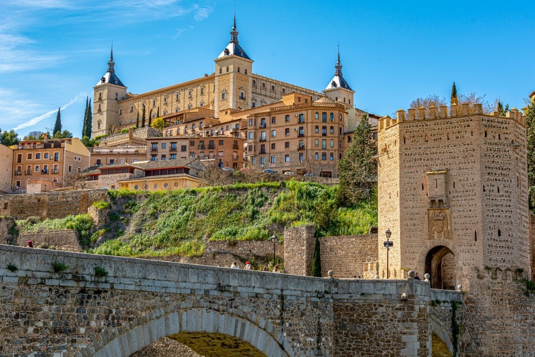 The beige city of Toledo, Spain is in view with several blocky buildings with square and octagonal columns; a stone bridge is in the foreground.