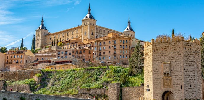 The beige city of Toledo, Spain is in view with several blocky buildings with square and octagonal columns; a stone bridge is in the foreground.