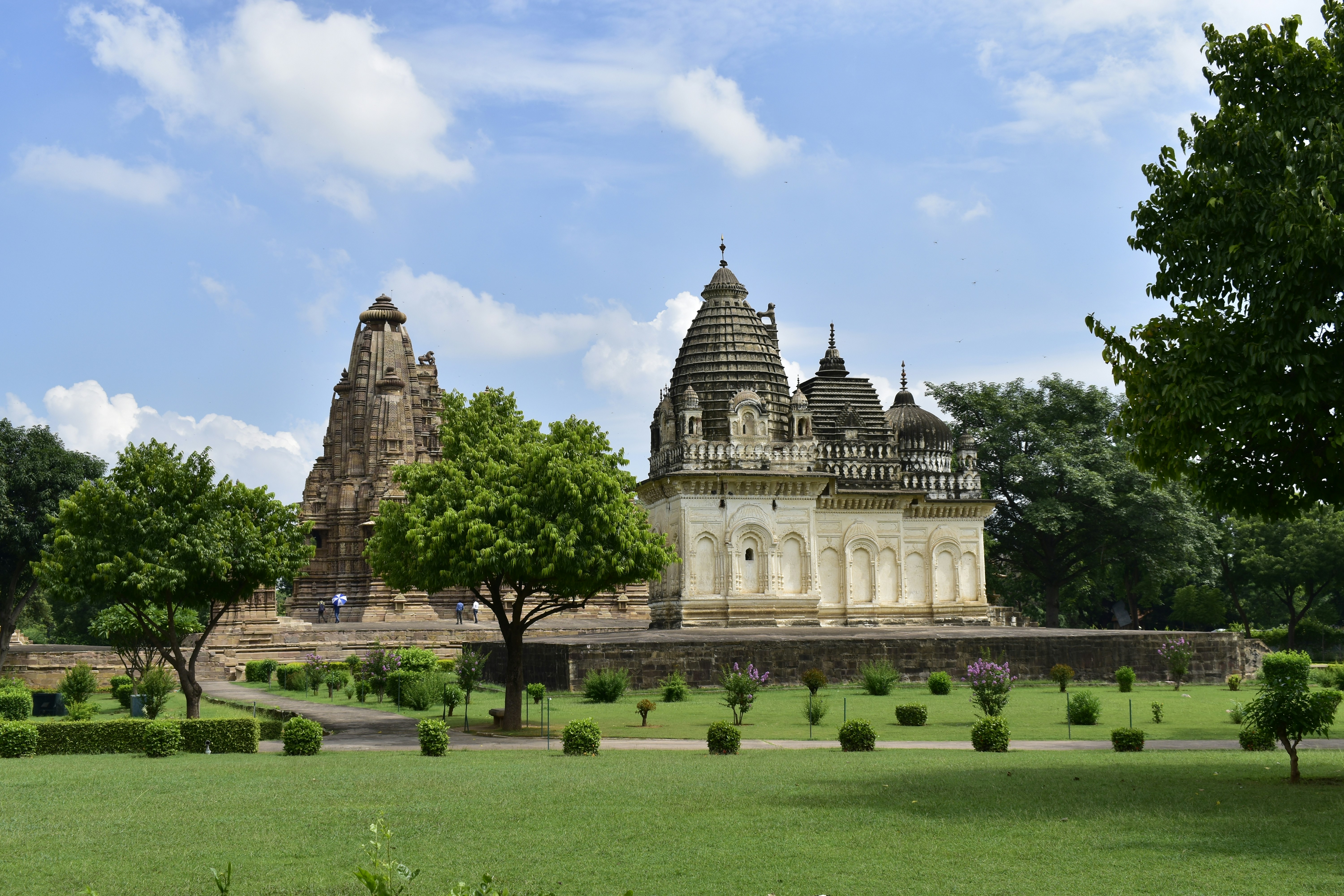 The Khajuraho Temple in India is shown from a distance, with a perfectly manicured, bright green garden in front of it and some luscious green trees to the right, behind it, and a couple to the left. The sky is partially cloudy.